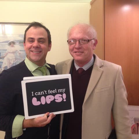 A smiling patient holding a humorous sign about local anaesthesia alongside a practitioner